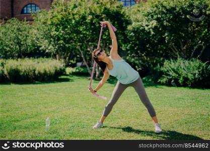 Full length shot of active woman stretches arms with fitness gum has good flexibility wears t shirt leggings and sneakers has workout with resistance band poses outdoor on green lawn during summer