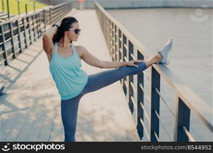 Full length shot of active woman stretches legs before run leans at fence dressed in sportsclothes sneakers sunglasses poses outdoor has regular training goes in for sport every day. Warming up