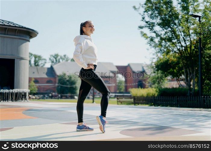 Full length shot of athletic sporty woman has rest after aerobics pilates workout enjoys sport activities outdoors wears white sweatshirt leggings and sneakers listens music keeps hands on waist
