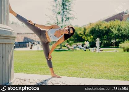 Full length shot of brunette woman stretches legs shows good flexibility does splits dressed in activewear sunglasses poses outside leads active lifestyle. People sport and aerobics concept.