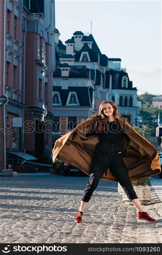 Full length shot of cute happy female model in fashionable black jumpsuit and yellow coat jumping in the street, city background, spring or autumn. Motion blur image. Fun lifestyle happiness concept