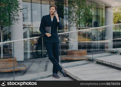 Full length shot of elegant businessman in formal clothes has telephone conversation holds digital gadget under arm answers phone call stands outdoor near office building or business center.. Full length shot of elegant businessman in formal clothes has telephone conversation