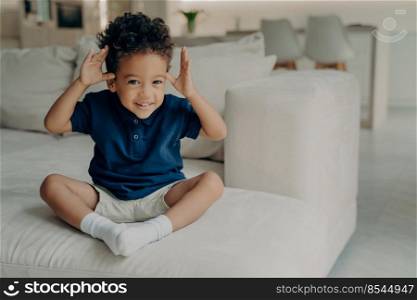 Full length shot of happy playful curly mulatto child holding his hands up, making funny face expression while sitting alone on couch, spending carefree and leisure time at home. Childhood concept. Playful little mulatto boy with hands up looking with excited expression at camera