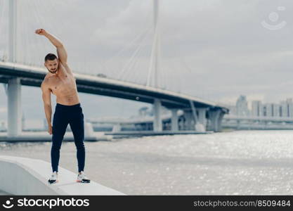 Full length shot of muscles active man does sport exercises outdoor poses near river bridge wears trousers and sneakers has beautiful male torso achieves best shape. Healthy lifestyle concept