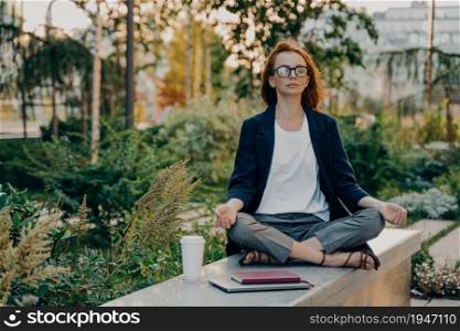 Full length shot of relaxed ginger woman meditates sits in lotus pose wears formal clothes keeps legs crossed breathes deeply spends free time in park surrounded by takeaway coffee tablet and notepad. Full length shot of relaxed ginger woman meditates sits in lotus pose wears formal clothes