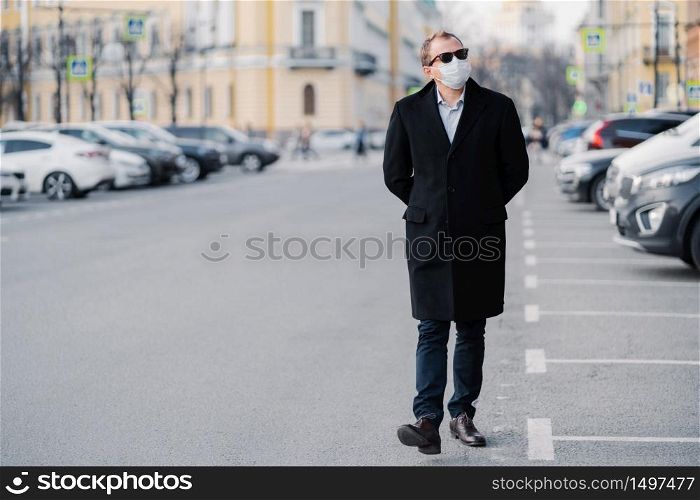 Full length shot of serious man walks at street, dressed in elegant outfit, wears medical mask to prevent coronavirus or another type of virus, stays safe during quarantine time. Pandemic situation