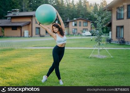 Full length shot of serious slim brunette woman has perfect body shape holds fitness ball over head, does fitness exercises outdoor near house, dressed in cropped top, leggings and sneakers.