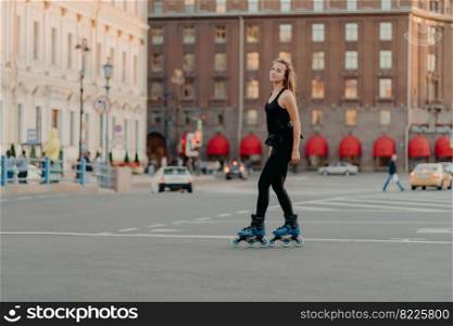 Full length shot of slim dark haired woman rollerblades on asphalt road dresed in black sportsclothes enjoys outdoor activities exercises for healthy and strong body. Sport and hobby concept