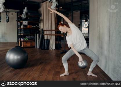 Full length shot of sportive red haired woman doing plie, holding in one hand small fitball while performing barre workout in studio or gym with sport equipment ambiance. Fitness activities concept. Slim young red haired woman ballerina doing plie with small fitball in hand