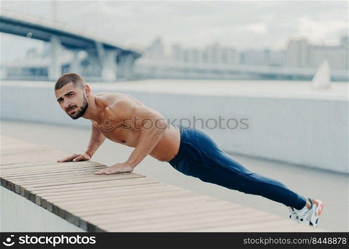 Full length shot of sporty muscular man in sportswear does push up exercise has concentrated serious expression poses outdoor. Self determined bearded sportsman planking for having strong arms