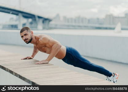 Full length shot of sporty muscular man in sportswear does push up exercise has concentrated serious expression poses outdoor. Self determined bearded sportsman planking for having strong arms