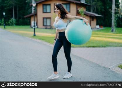 Full length shot of sporty slim woman holds big inflated fit ball, dressed in active wear, has aerobics exercises outdoors, has athletic body stands on road with house in background. People, lifestyle