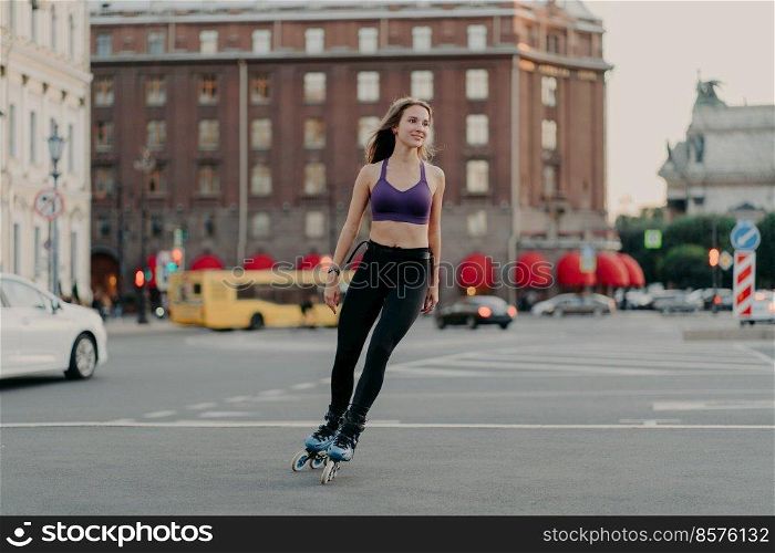 Full length shot of sporty slim woman in active wear rides on blades enjoys outdoor fitness activity during warm summer day poses at urban place on asphalt. Rollerblading and leisure concept