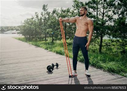 Full length shot of strong bearded sportsman flexes muscles with elastic resistance band, wears sport trousers and sneakers, stands outdoor, uses sport equipment for workout, performs exercises