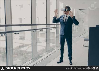 Full length shot of successful handsome business professional in elegant suit standing in office and using VR technologies at work, engaged in virtual conference with colleagues, wearing 3d goggles. Successfu business professional in elegant suit standing in office and using VR technologies at work