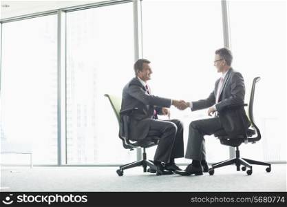 Full-length side view of businessmen shaking hands while sitting on office chairs by window