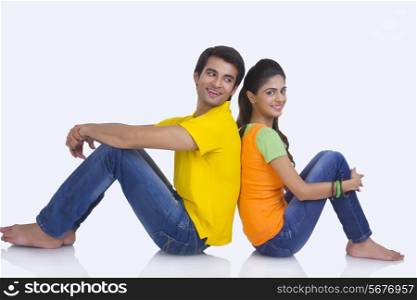 Full length side view of happy young couple sitting back to back on white background