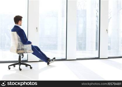Full length side view of thoughtful young businessman sitting on chair by windows in new office