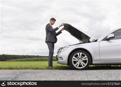 Full length side view of young businessman opening broken down car hood at countryside