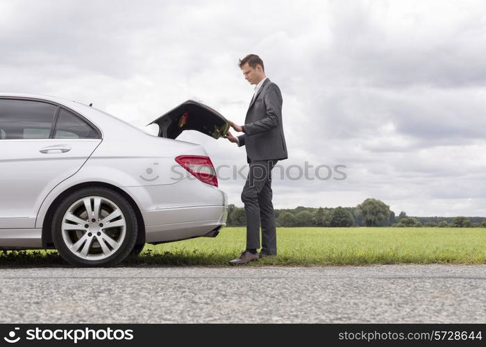 Full length side view of young businessman opening broken down car trunk at countryside