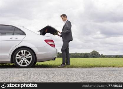 Full length side view of young businessman opening broken down car trunk at countryside