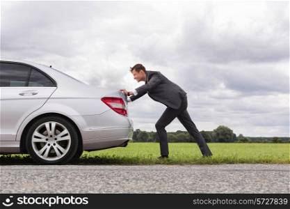 Full length side view of young businessman pushing broken down car on road