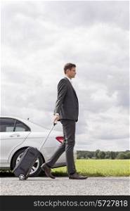Full length side view of young businessman with luggage walking by broken down car at countryside