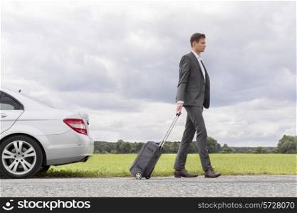 Full length side view of young businessman with suitcase leaving broken down car at countryside