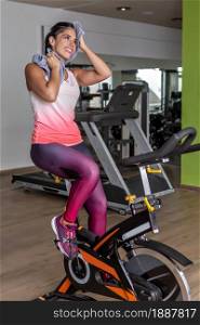 Full length view of a young sportswoman looking away doing exercise in bike while wiping her face with a towel at gym. Full length view of a young sportswoman doing exercise in bike while wiping her face with a towel at gym