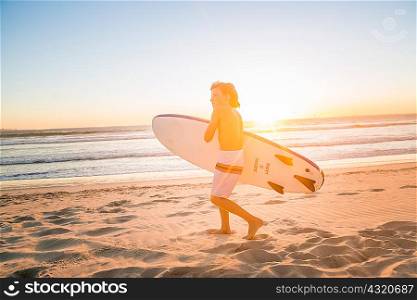 Full length view of boy carrying surfboard on beach at sunset