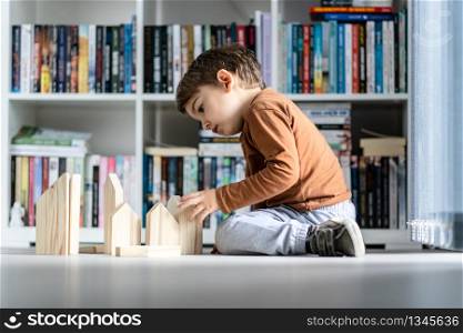 full length view of small caucasian boy little child ar home kid playing with wooden toys brick in shape of house sitting on the wooden or vinyl laminated floor alone developing creativity side view