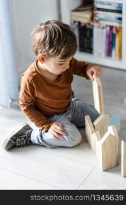 full length view of small caucasian boy little child ar home kid playing with wooden toys brick in shape of house sitting on the wooden or vinyl laminated floor alone developing creativity top view