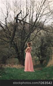 full length young stylish woman near blossoming tree in the spring park. blonde girl with hairstyle in pink dress.. full length young stylish woman near blossoming tree in the spring park. blonde girl with hairstyle in pink dress