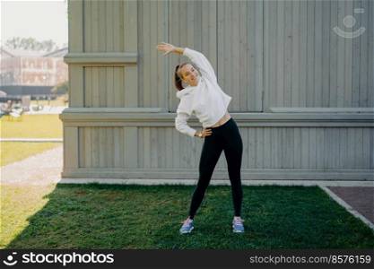 Full≤n>h shot of active sportswoman does exercises outdoors stetches body≤ans at different sides wears sweatshirt trousers and trai≠rs listensμsic in earpho≠s has fir body smi≤s hapπly