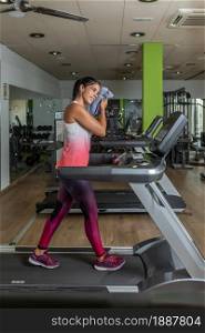 Full vertical image of a young latin adult sportswoman wiping face with towel while walking on a treadmill at gym.. Vertical image of a young adult sportswoman wiping face with towel while walking on a treadmill at gym