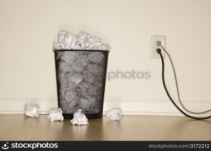 Full wire mesh trash can with crumpled paper next to electrical outlet and plugs.