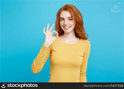 Fun and People Concept - Headshot Portrait of charming ginger red hair girl with freckles smiling and making ok sign with finger. Pastel blue background. Copy Space.