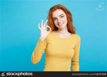 Fun and People Concept - Headshot Portrait of charming ginger red hair girl with freckles smiling and making ok sign with finger. Pastel blue background. Copy Space.