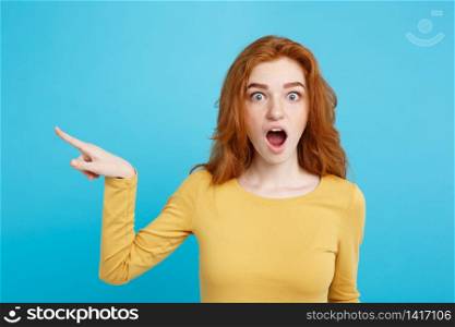 Fun and People Concept - Headshot Portrait of happy ginger red hair girl with pointing finger away and shocking expression. Pastel blue background. Copy Space.