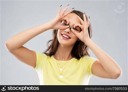 fun and people concept - smiling young woman or teenage girl looking through finger glasses over grey background. young woman looking through finger glasses