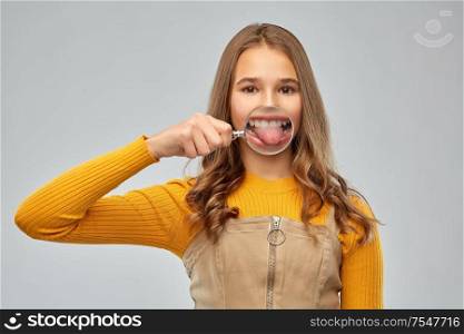 fun, dental care and people concept - happy teenage girl showing her teeth and tongue through magnifying glass and making silly faces over grey background. teenage girl shows tongue through magnifying glass