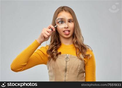 fun, discovery and vision concept - happy teenage girl looking through magnifying glass and making silly faces over grey background. teenage girl looking through magnifying glass