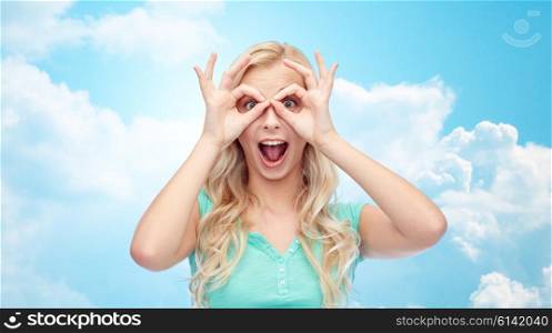 fun, emotions, expressions and people concept - smiling young woman or teenage girl looking through glasses made of fingers over blue sky and clouds background
