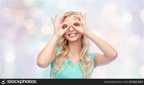 fun, emotions, expressions and people concept - smiling young woman or teenage girl looking through glasses made of fingers over holidays lights background
