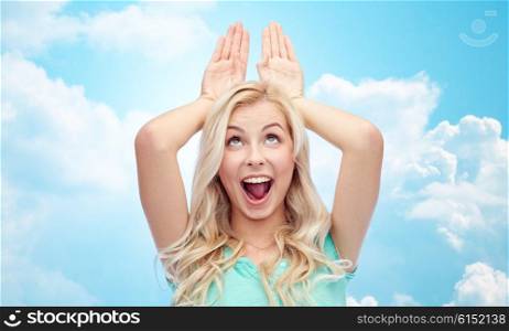 fun, expressions, easter and people concept - happy smiling young woman making bunny ears over blue sky and clouds background