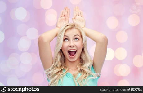 fun, expressions, easter and people concept - happy smiling young woman making bunny ears over pink holidays lights background