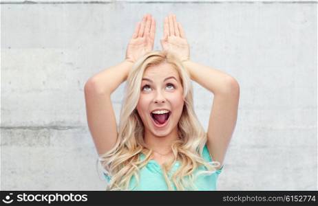 fun, expressions, easter and people concept - happy smiling young woman making bunny ears over gray concrete wall background