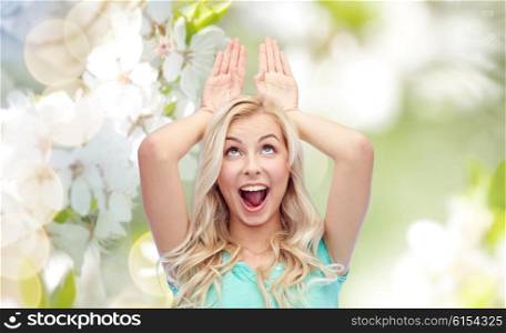 fun, expressions, easter and people concept - happy smiling young woman making bunny ears over natural spring background