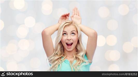 fun, expressions, easter and people concept - happy smiling young woman making bunny ears over holidays lights background