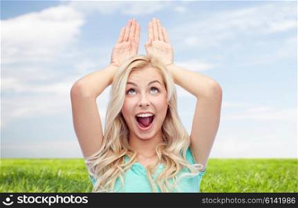 fun, expressions, easter, summer and people concept - happy smiling young woman making bunny ears over blue sky and grass background